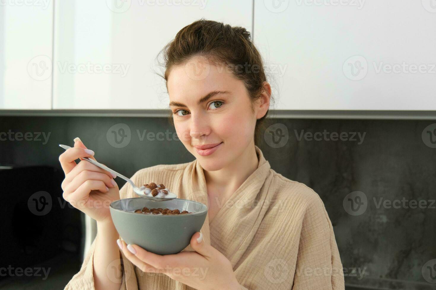 Good-looking brunette woman with bowl of cereals, eating meal for breakfast, wearing bathrobe, looking at camera and smiling photo