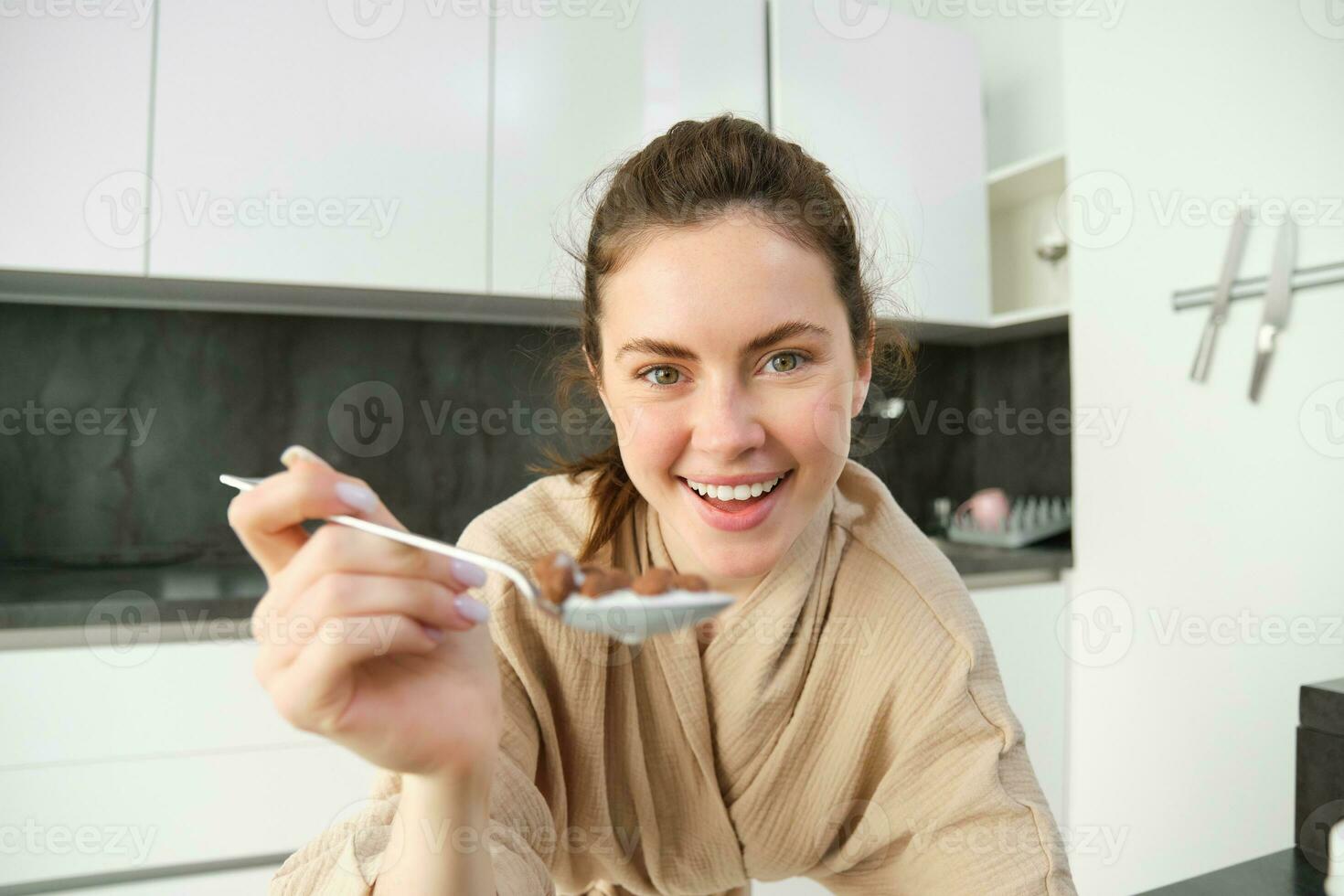 retrato de contento joven mujer se inclina en cocina encimera y comiendo cereales, tiene Leche y cuenco en frente de su, teniendo su desayuno, vistiendo bata de baño foto