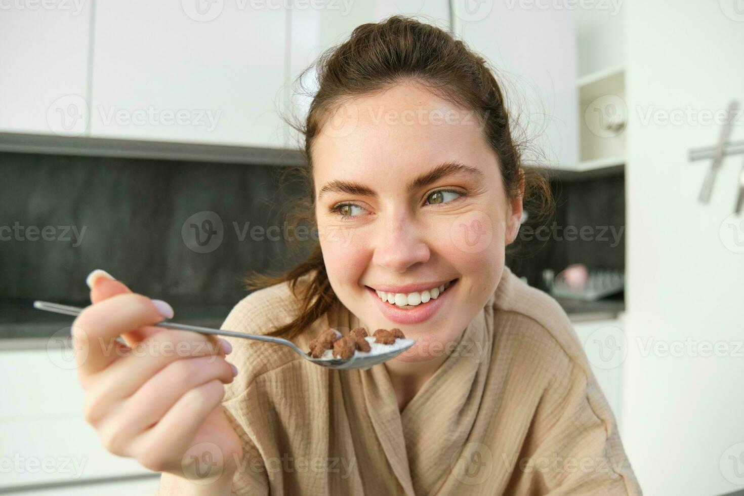 Image of happy young woman eating cereals at home with milk, having her breakfast, wearing bathrobe, sitting in kitchen alone photo