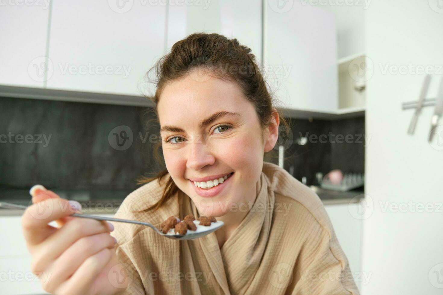 retrato de hermosa mujer a hogar, comiendo su desayuno, participación cuchara, propensión en encimera y teniendo cereales con Leche foto