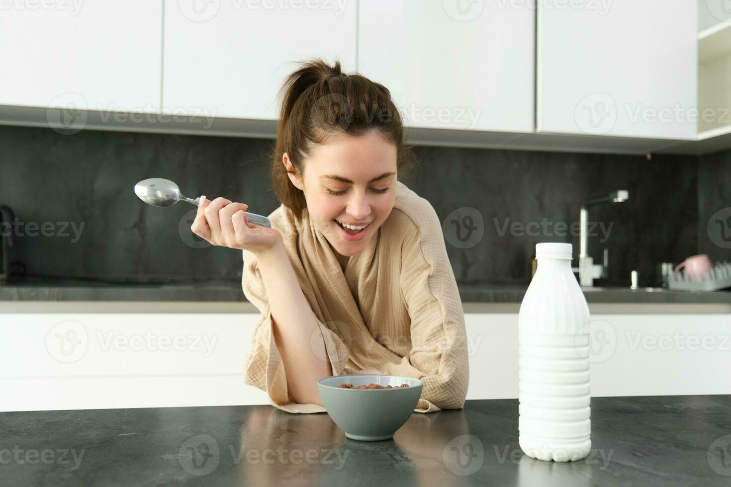 Portrait of young beautiful woman in bathrobe, eating cereals for breakfast, leans on kitchen worktop, looking at her morning meal photo