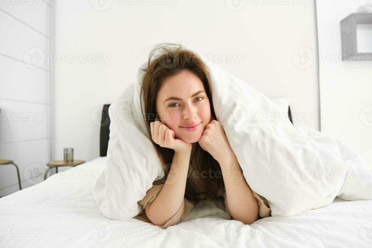 Portrait of attractive smiling woman relaxing in bed, lying covered in white sheets and warm duvet in morning, looking happy at camera photo