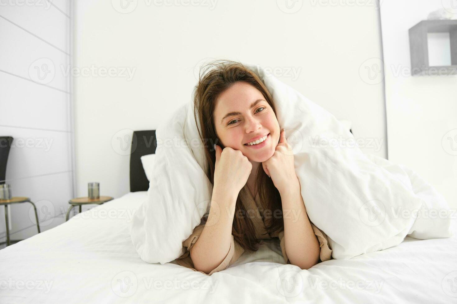 Portrait of gorgeous smiling woman, lying in bed covered in duvet, has messy hair, looking happy, relaxing in her bedroom, spending time in hotel room in morning photo