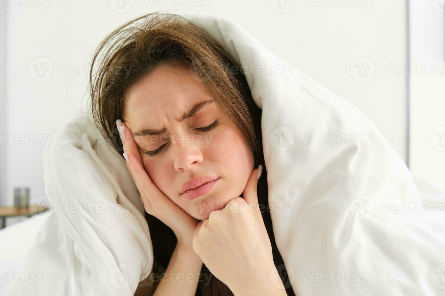 Woman feeling unwell in bed, lying in her bedroom under blanket and white sheets in morning, frowning and touching head, has headache or migraine photo