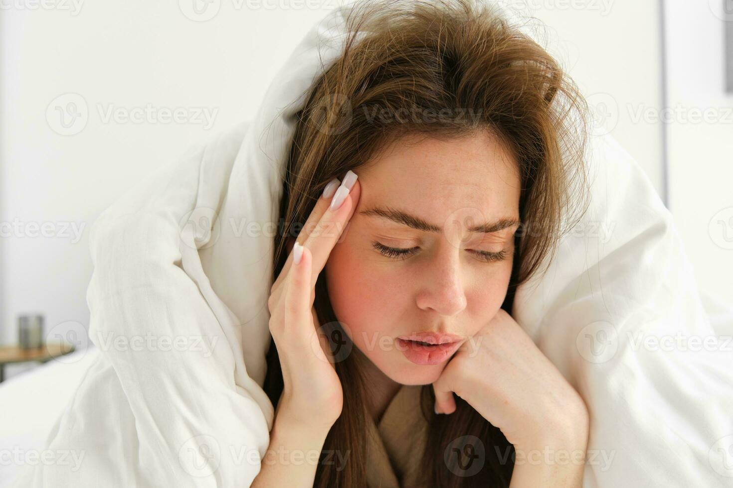 Portrait of sad woman feeling unwell, lying in bed under white blanket, touching head, has headache after hangover, has depression photo