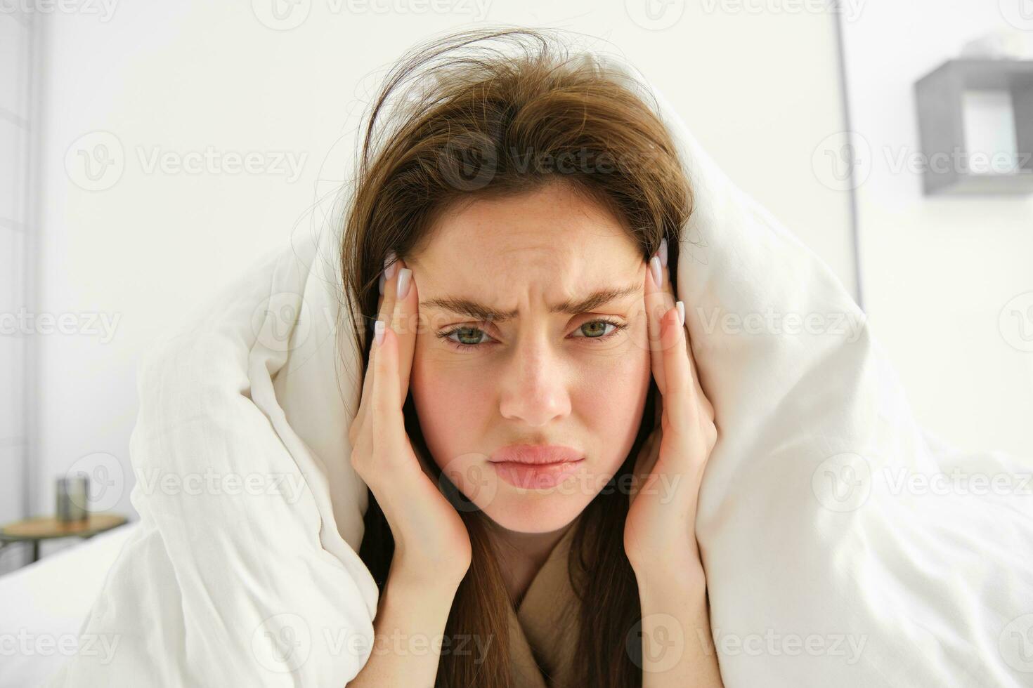 Woman feeling unwell in bed, lying in her bedroom under blanket and white sheets in morning, frowning and touching head, has headache or migraine photo