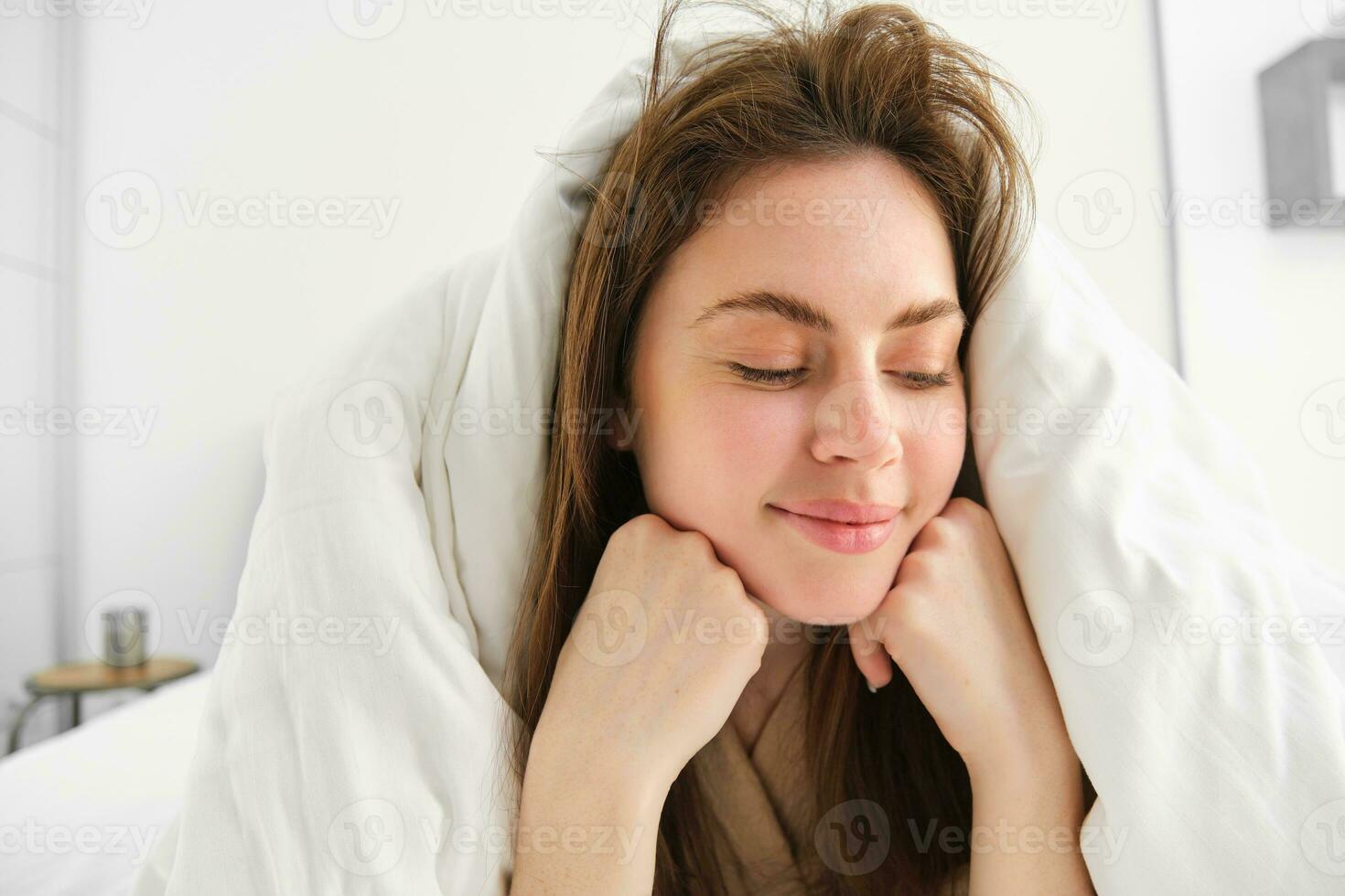 Cheerful woman feeling comfortable in bed, lying in bedroom covered in white sheets, smiling pleased, has messy hair in morning, waking up and looking happy photo