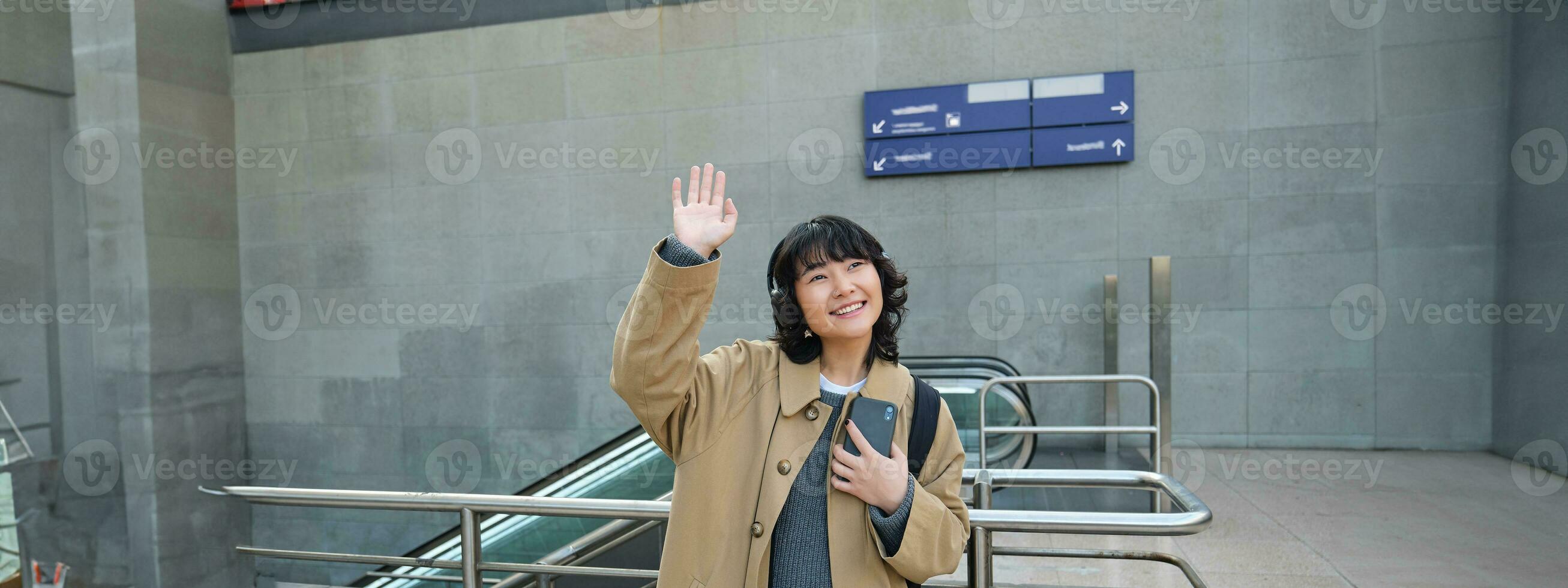 Portrait of cute smiling asian girl in headphones, stands on street with smartphone, waves hand at someone, says hello, greets friend photo