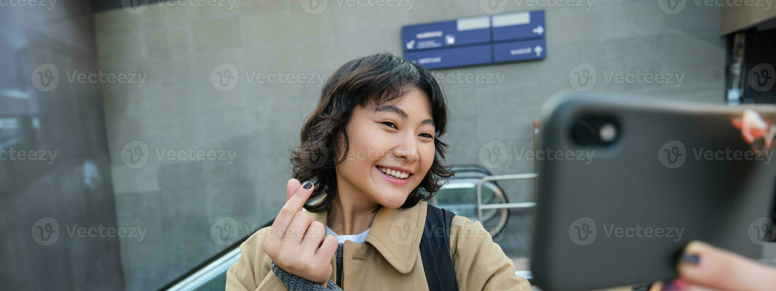 retrato de elegante coreano chica, estudiante toma selfie en calle, sostiene teléfono inteligente y poses en sus cámara, hace vídeo en turista viaje, sonrisas con alegría foto