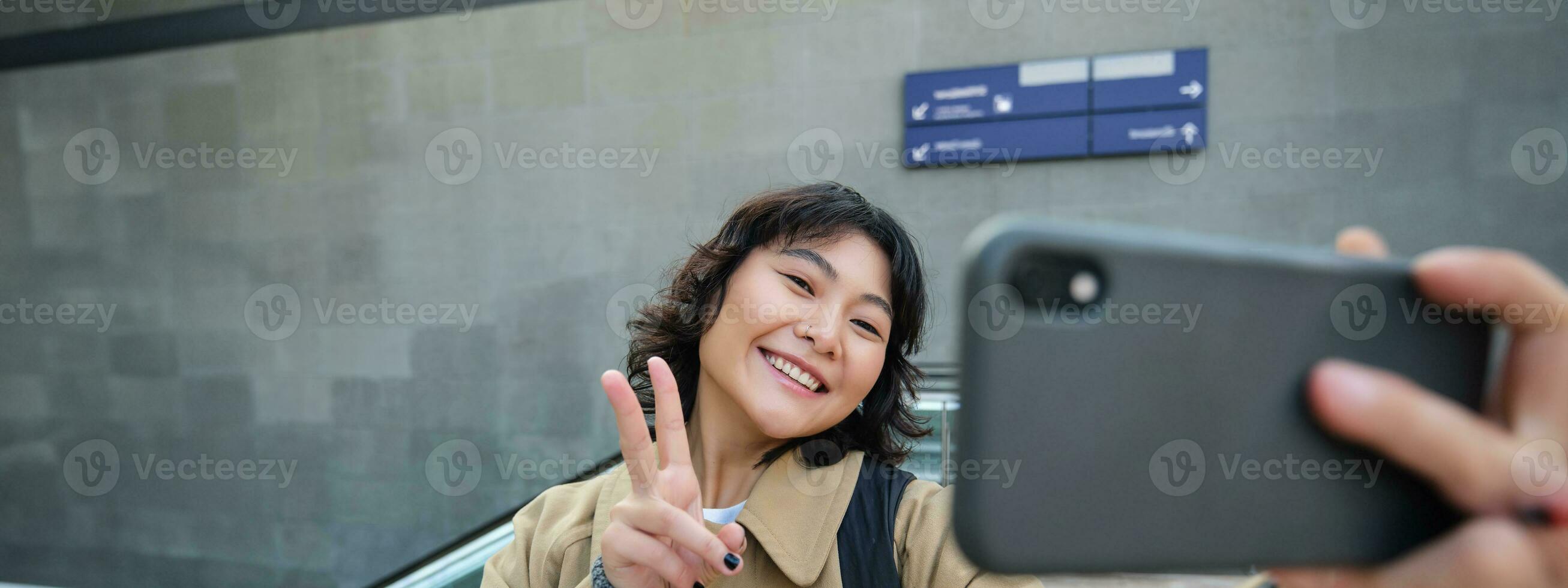 retrato de elegante coreano chica, estudiante toma selfie en calle, sostiene teléfono inteligente y poses en sus cámara, hace vídeo en turista viaje, sonrisas con alegría foto