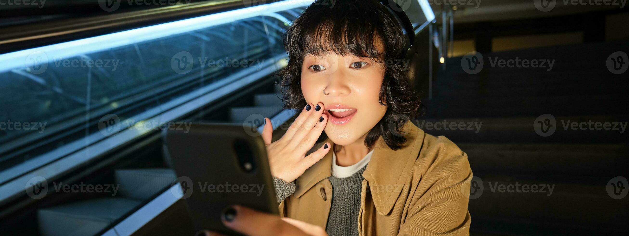 Close up portrait of korean girl looks surprised at mobile phone, reads something amazing on smarpthone screen, watching video in headphones, sits on staircase photo