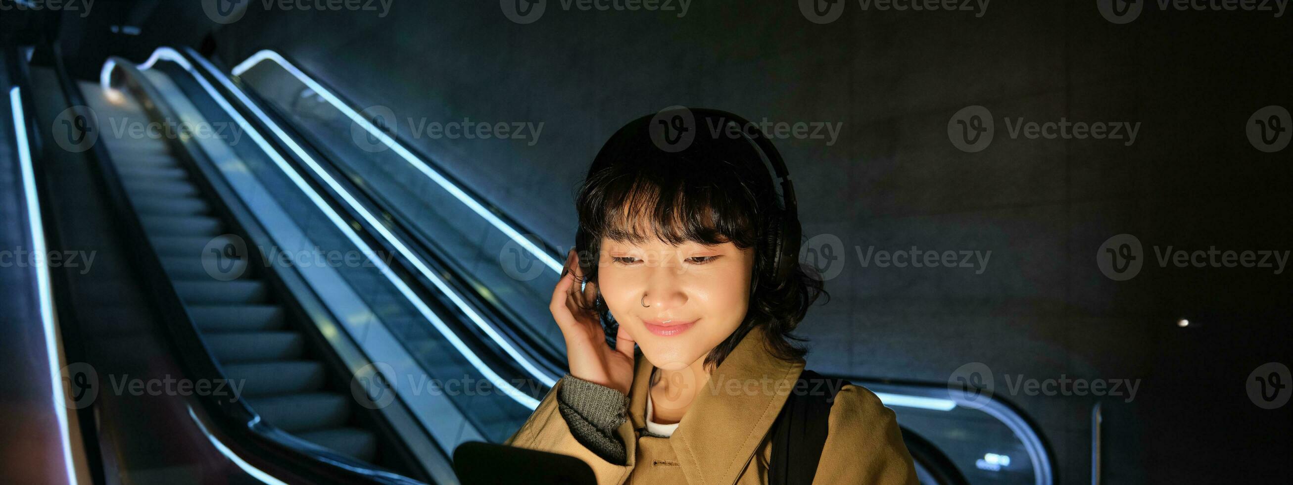 Cute young student, girl in headphones, commuting, standing near escalator, holding smartphone, listening music or podcast while travelling in city photo