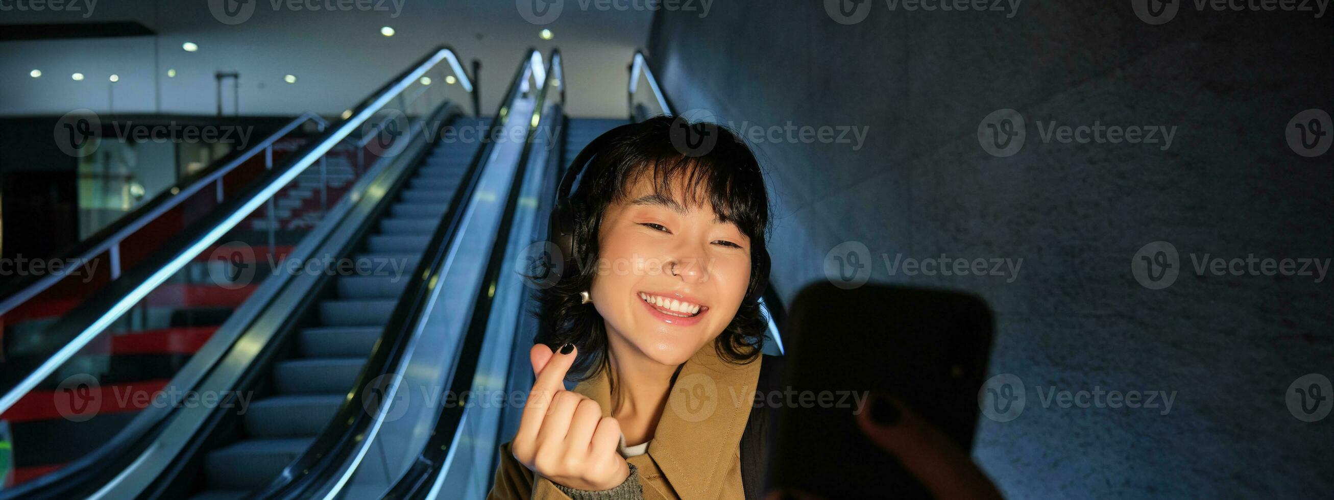 elegante sonriente asiático chica, tomando selfie en móvil teléfono mientras montando escalera mecánica, yendo abajo a metro estación, demostración dedo corazón y posando para foto