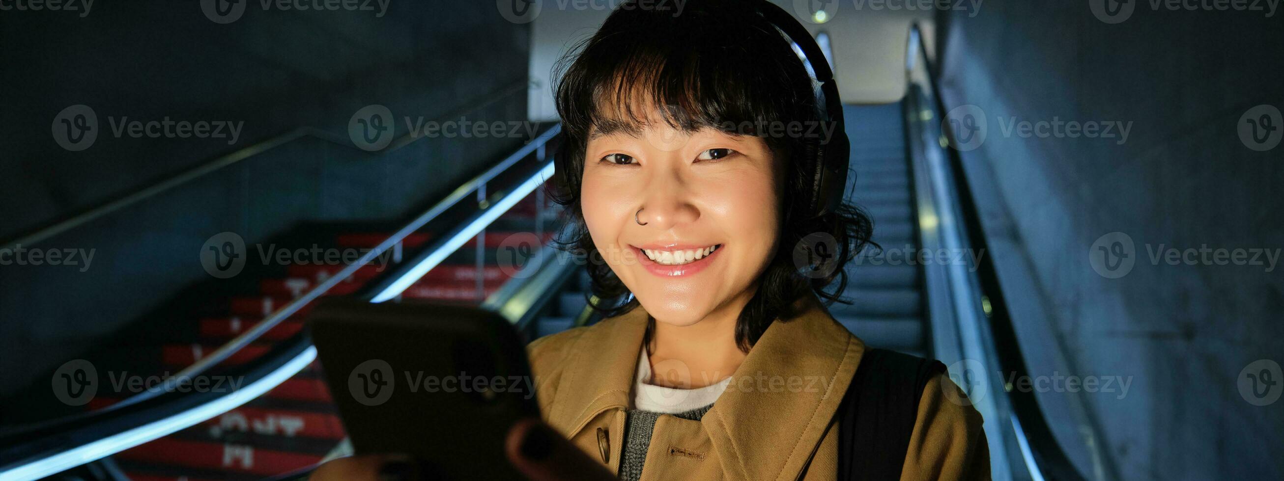 Close up of cute asian girl in headphones, picks song to listen while commuting, going down escalator to metro, smiling as reading message on mobile phone photo