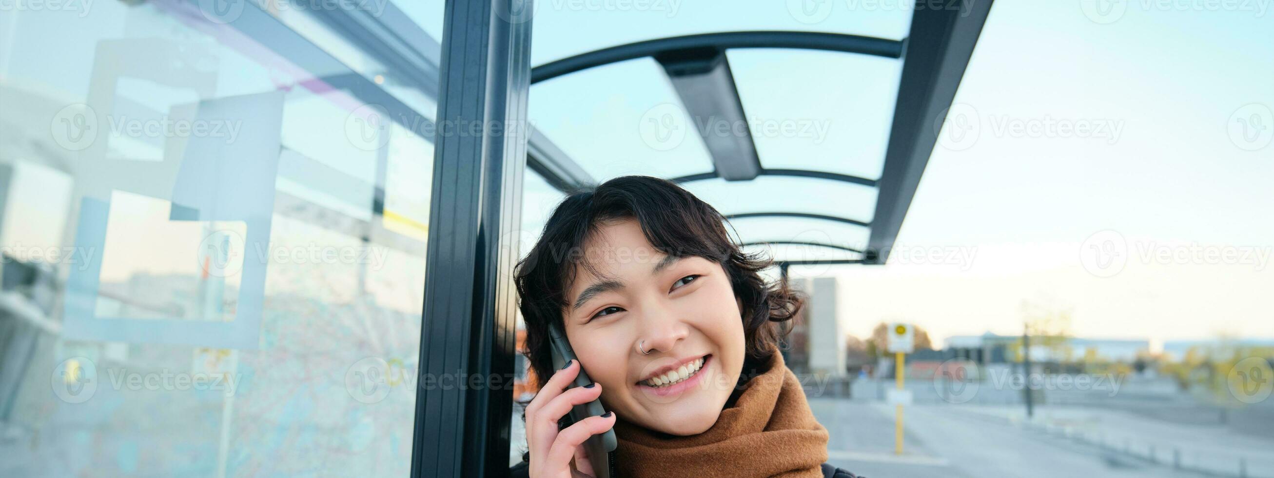 Close up of cute Korean woman, making a phone call, talking and smiling on telephone, standing in winter jacket on bus stop, waiting for her transport to arrive photo