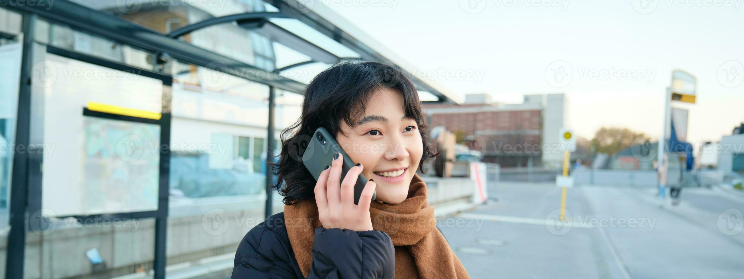 Smilling Korean girl talking on mobile phone, standing on bus stop, using smartphone, posing on road in winter, wrapped in scarf, wearing black jacket photo