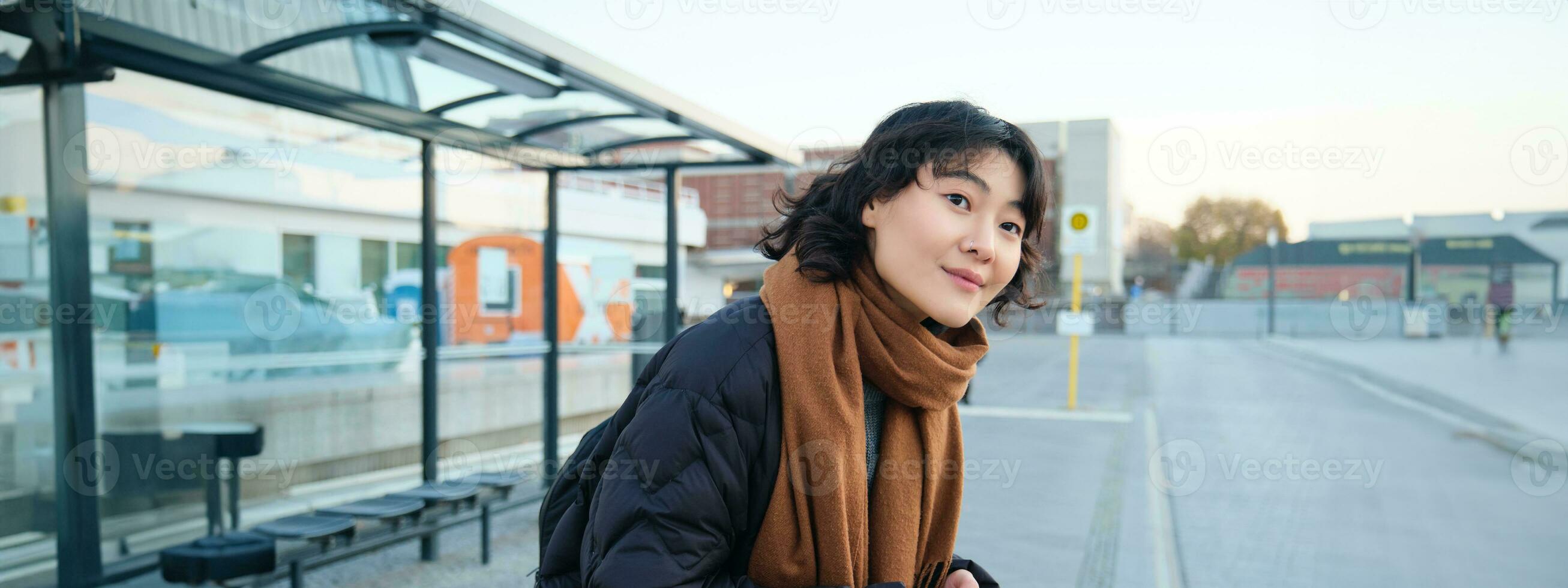 Cute smiling woman standing on a bus stop and looking at road, waiting for her public transport, holding smartphone, checking schedule on mobile app photo