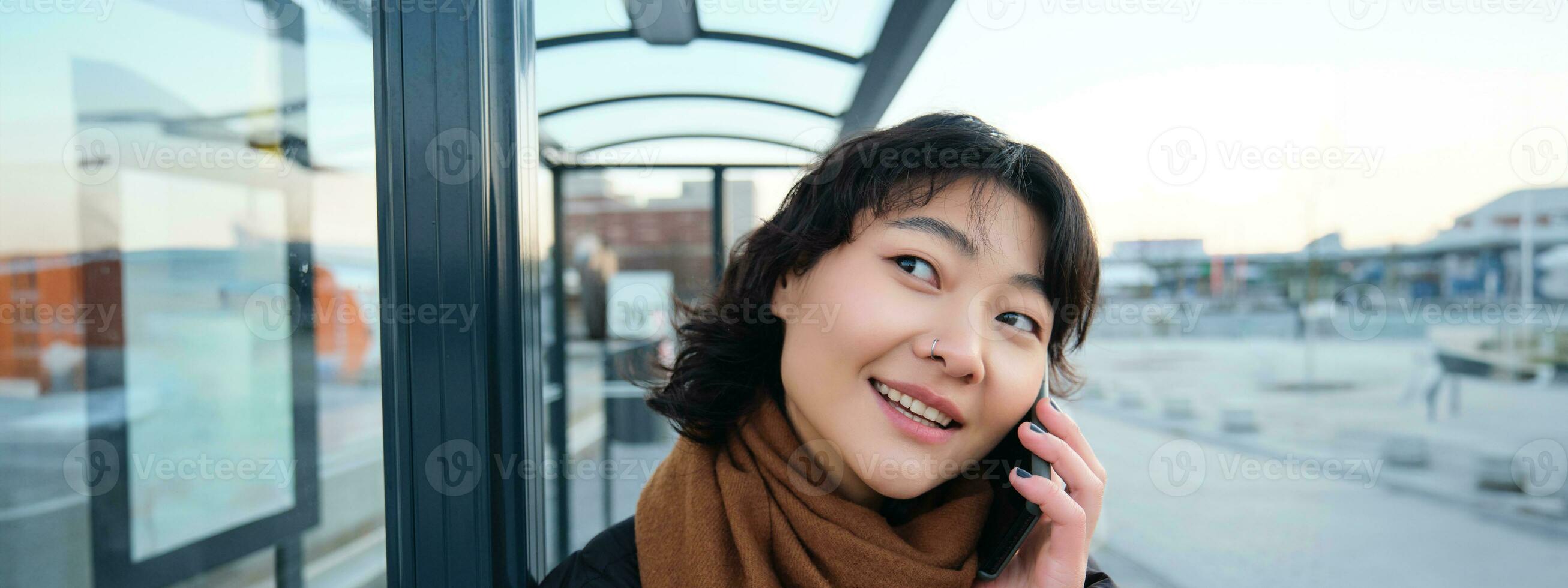 Close up of cute Korean woman, making a phone call, talking and smiling on telephone, standing in winter jacket on bus stop, waiting for her transport to arrive photo