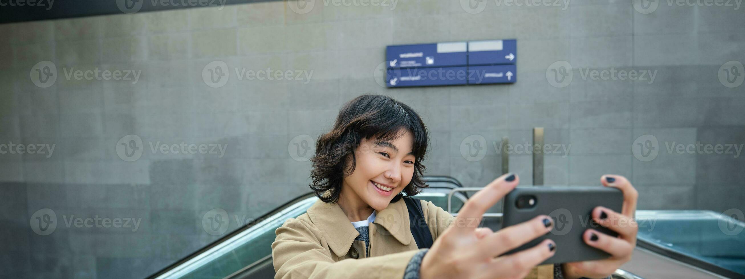 Portrait of stylish korean girl, student takes selfie on street, holds smartphone and poses on its camera, makes video on tourist trip, smiles with joy photo