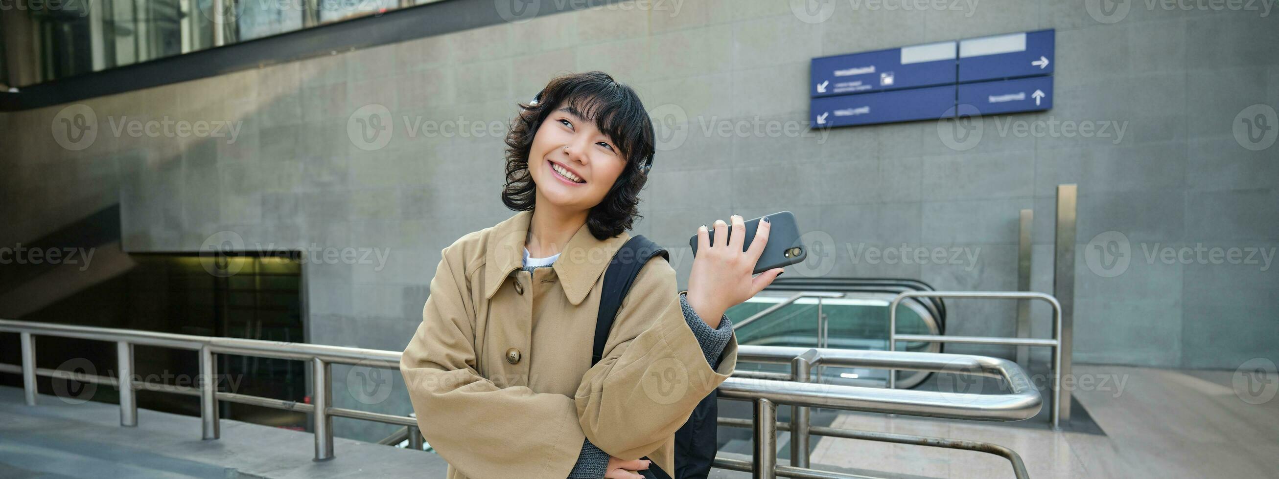 Happy young woman in headphones, stands on street with smartphone and drinks coffee, waits for commute train or bus, travels in city photo