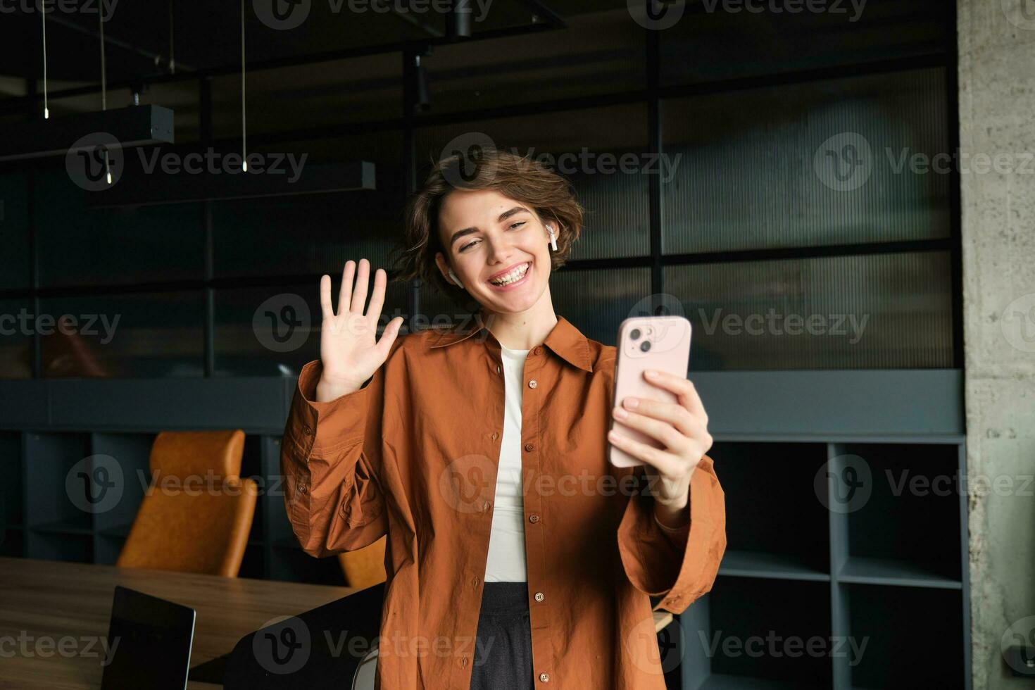 Happy young woman, office employee in casual clothes, waves hand at smartphone and says hello to coworkers joining online meeting, video chats, smiles at screen photo