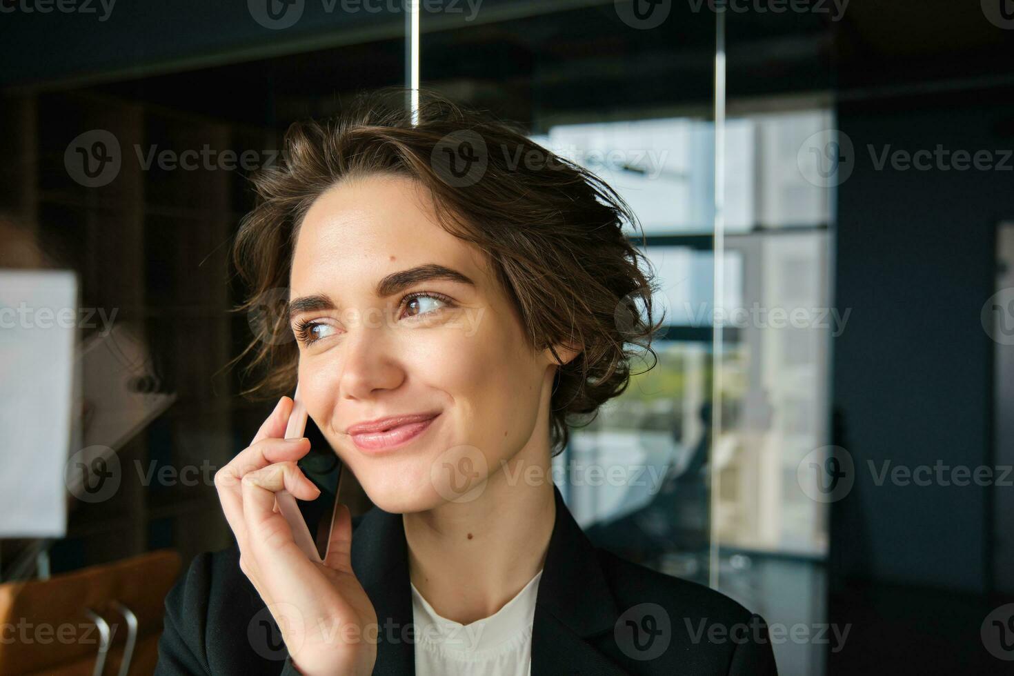 Image of confident businesswoman answering phone call, having a conversation over the telephone and smiling, standing in office or co-working space photo