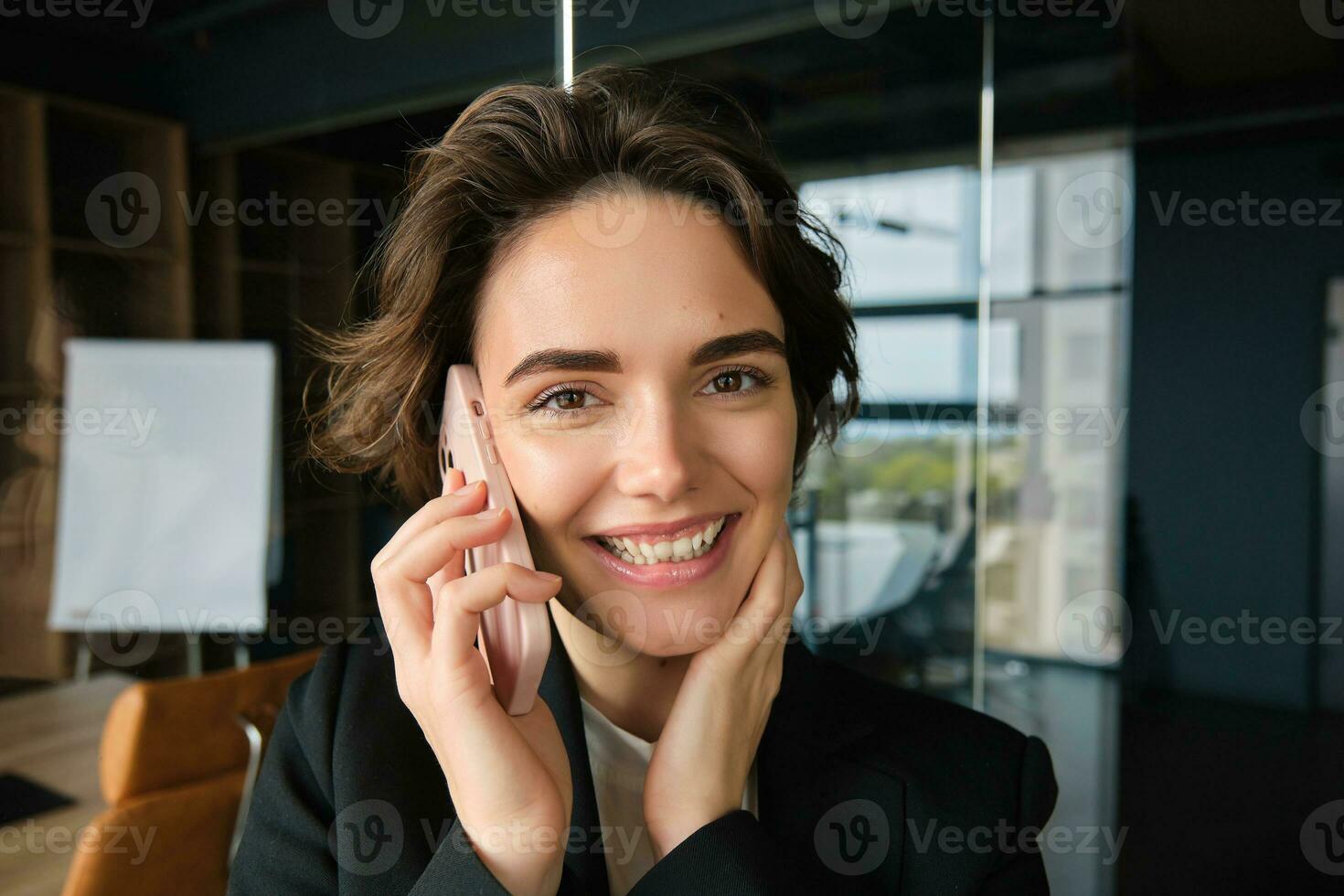 Close up portrait of smiling saleswoman, office manager stands in conference room, answers phone call, talking on mobile telephone with someone, receives great news and rejoices photo