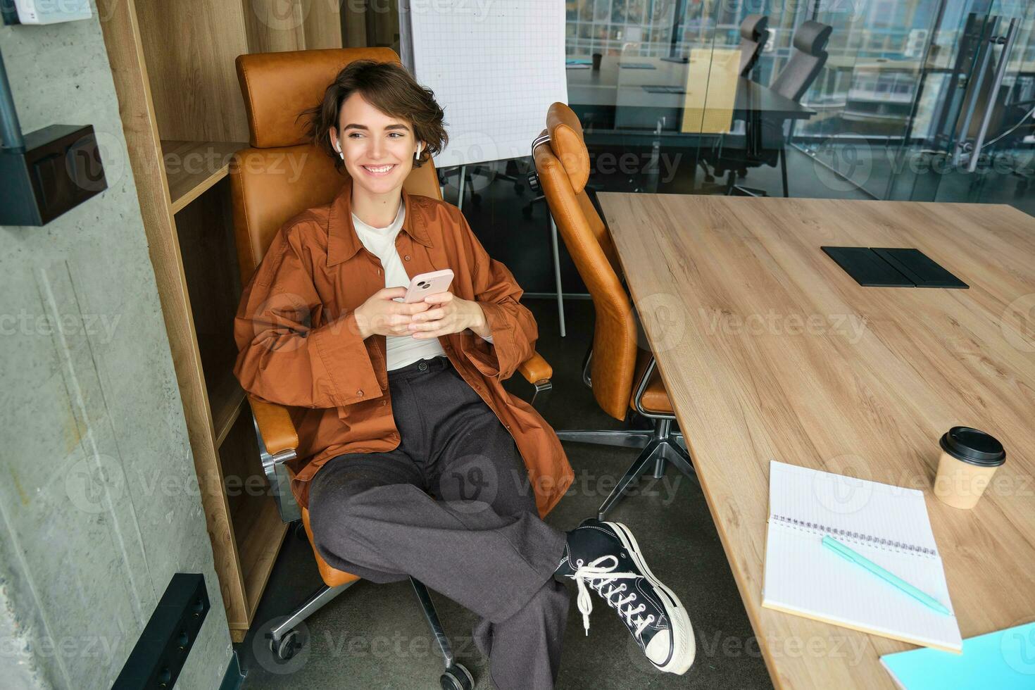 imagen de joven profesional emprendedor, mujer en un oficina, sentado en silla en un oficina, utilizando móvil teléfono, mensajería un compañero de trabajo foto