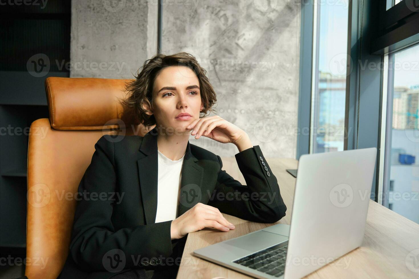 Portrait of confident businesswoman in office, sitting with laptop, looking serious outside window, thinking, solving issue at work photo