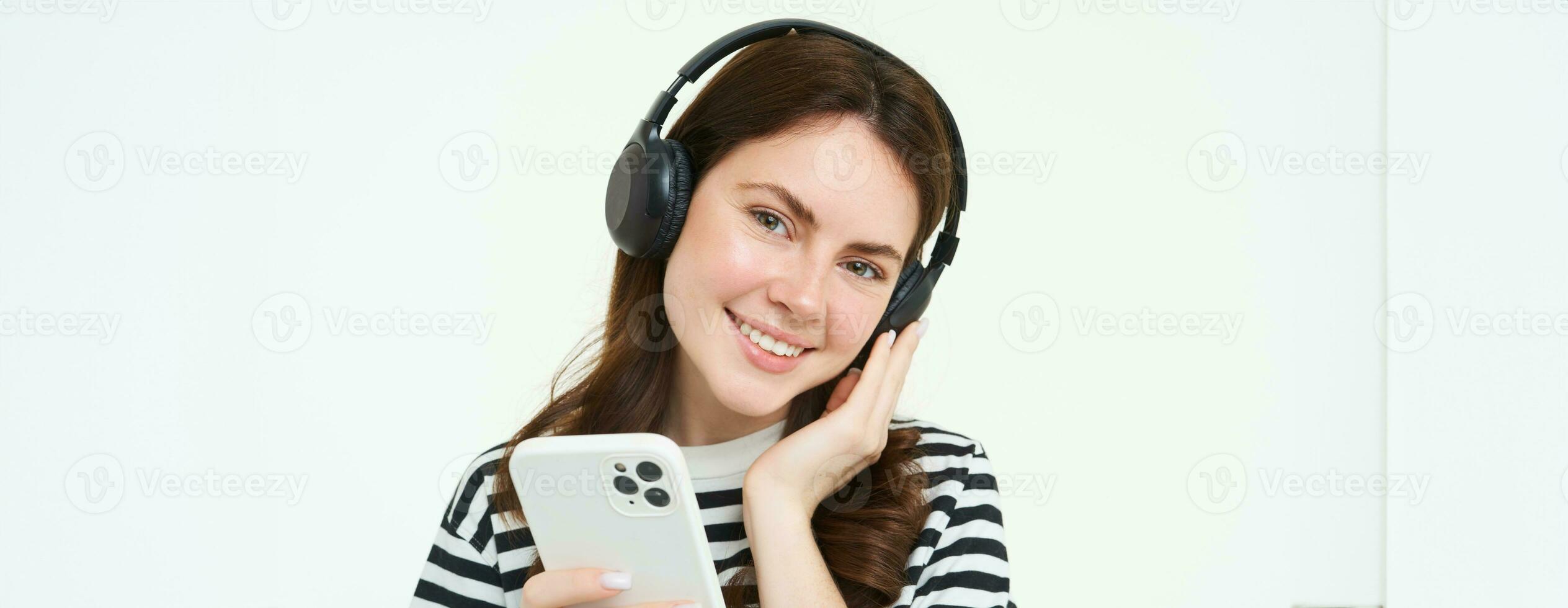 Smiling, beautiful female student in headphones, holding mobile phone, smiling and listening music, using smartphone, standing over white background photo