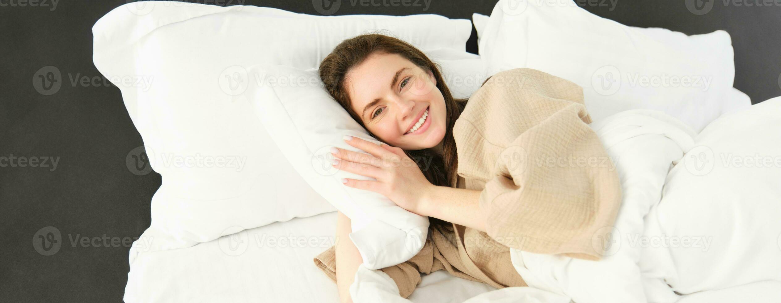 Gorgeous woman resting in hotel room, sleeping in bed, lying on pillow with eyes opened and smiling, having lazy morning photo