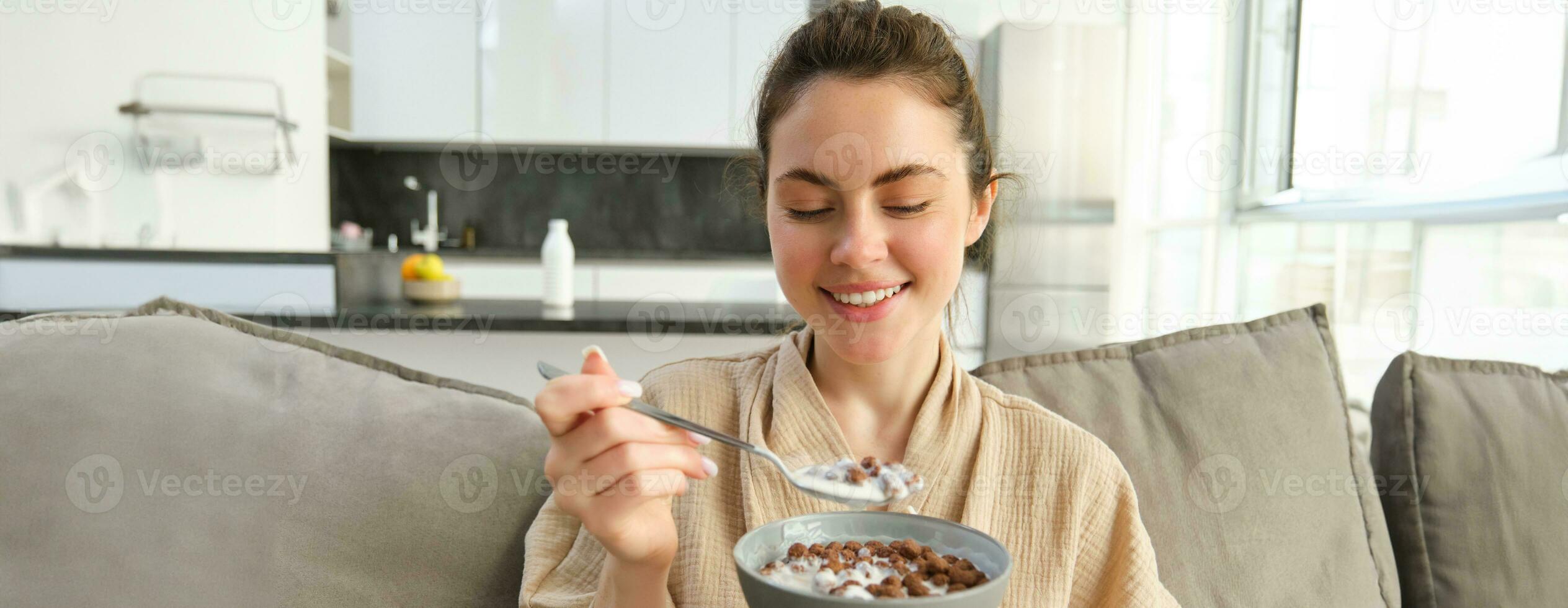 Portrait of gorgeous brunette woman sits on sofa, having her breakfast at home, smiling while eating cereals, holding bowl and spoon and laughing photo