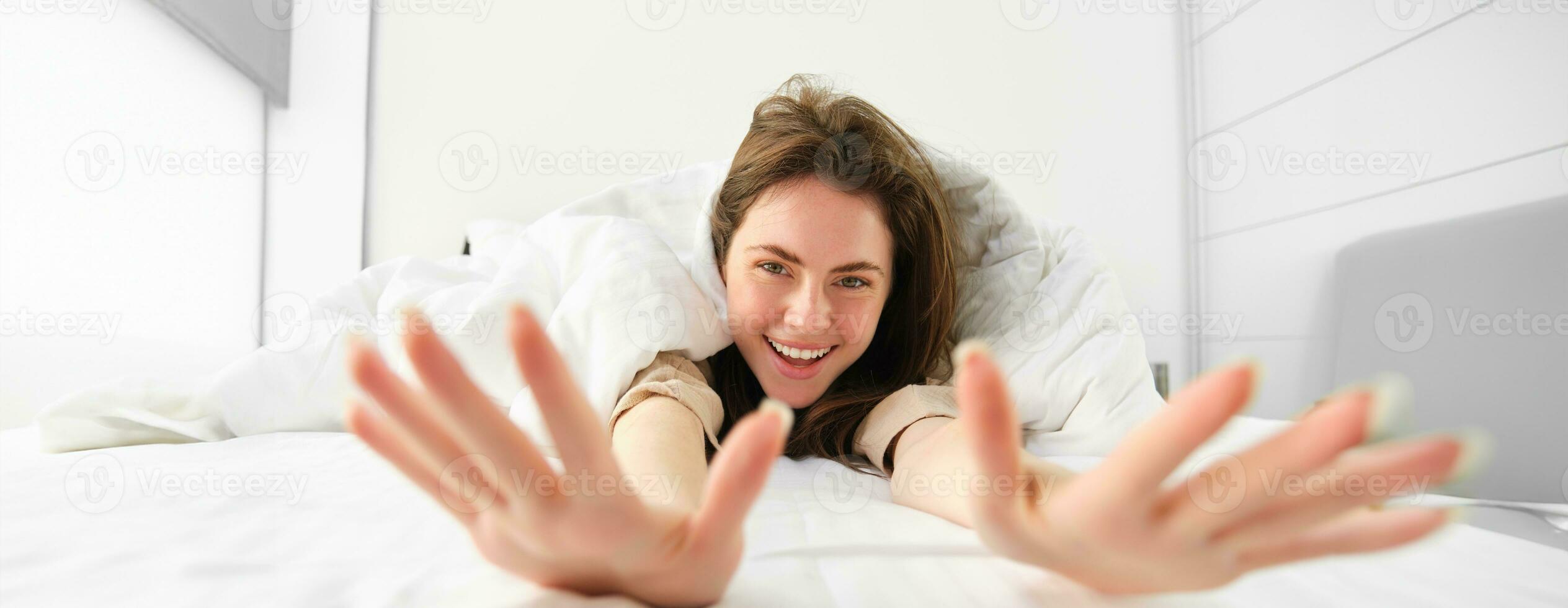 Beautiful woman stretching, showing her arms into camera, lying in bed, waking up in morning from good sleep or nap photo