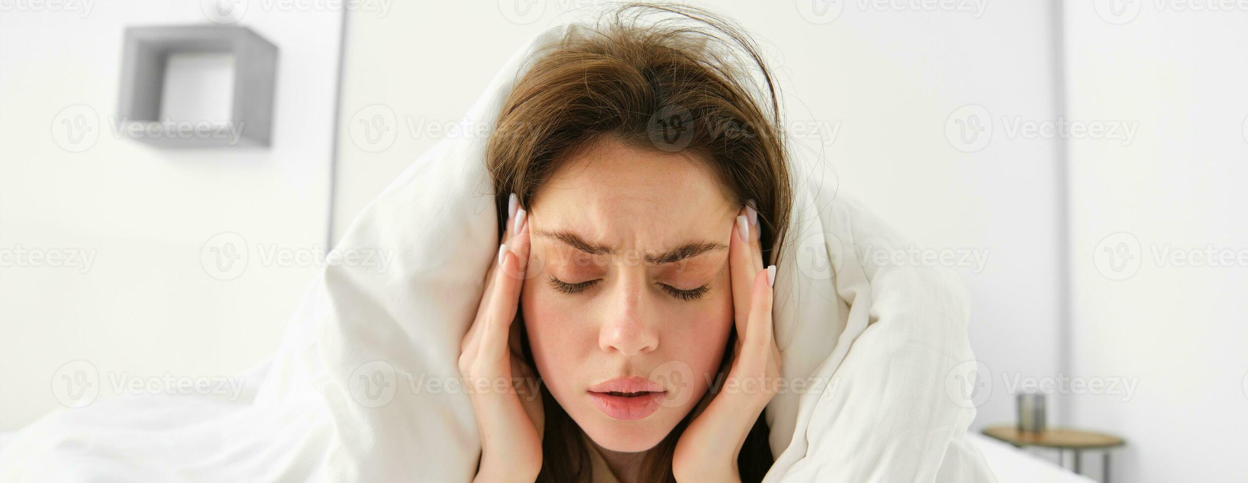 Close up of woman feeling unwell at home, lying in bed under white blankets and frowning, touching head, has headache, migraine, recovering from covid photo