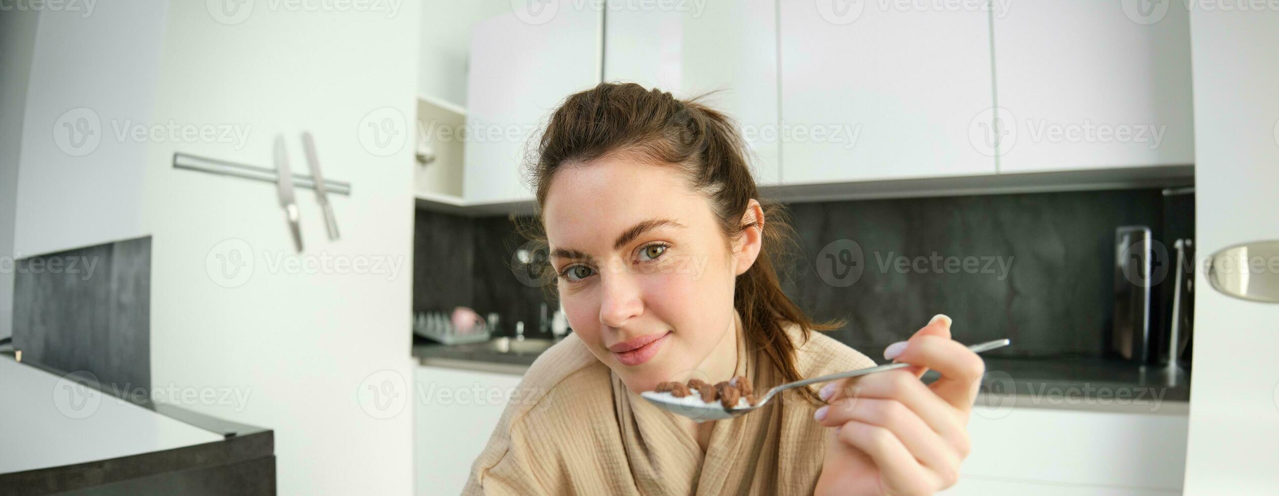 cerca arriba retrato de contento sonriente morena en bata de baño, participación cereales en cuchara, comiendo desayuno con Leche y mirando alegre, comenzando su día en Mañana foto