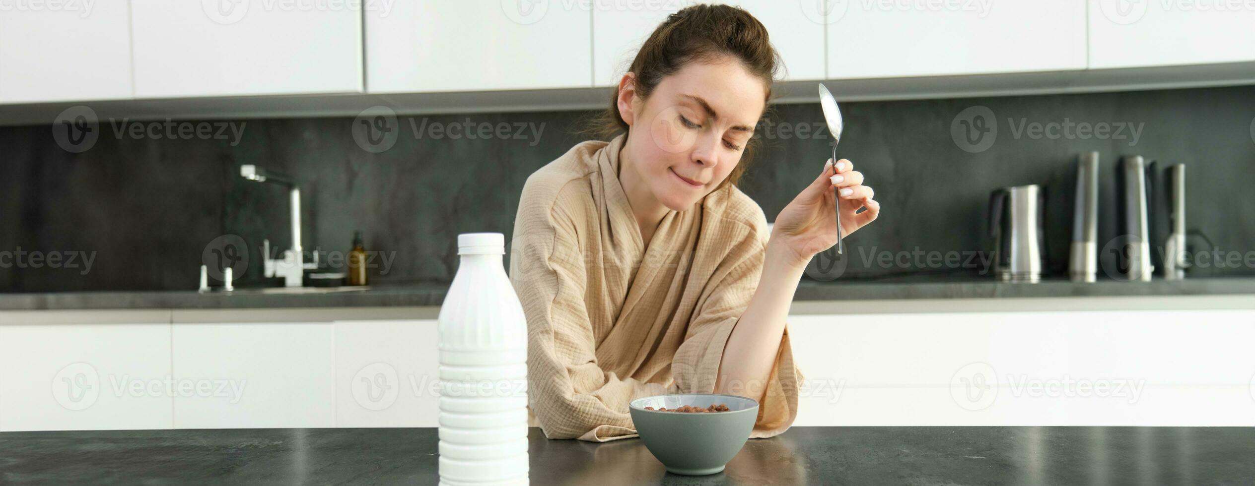 retrato de contento joven mujer se inclina en cocina encimera y comiendo cereales, tiene Leche y cuenco en frente de su, teniendo su desayuno, vistiendo bata de baño foto