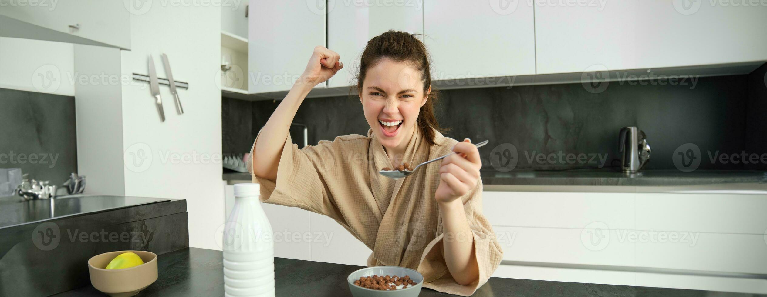 Portrait of enthusiastic young woman eating cereals with milk, looking excited and happy, sitting near kitchen worktop and having breakfast, raising hand up in triumph photo