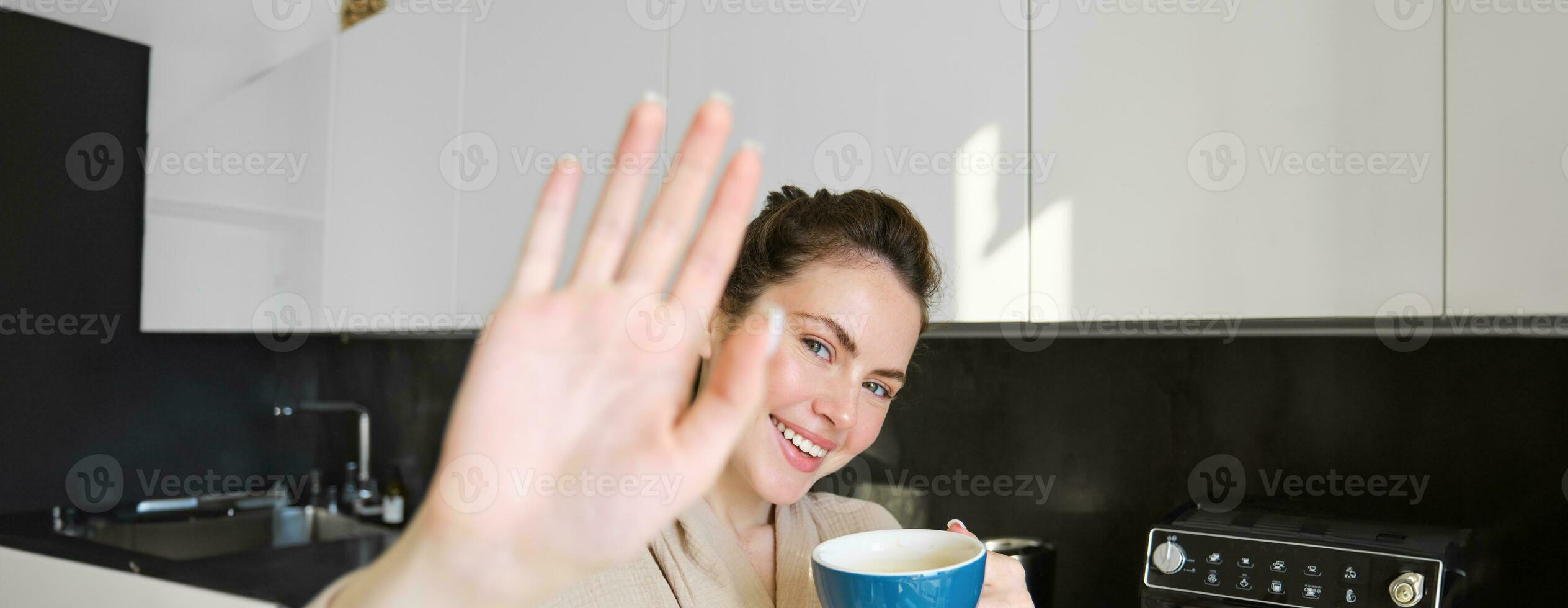 Portrait of cute young woman, covers herself from camera, laughing and flirting, drinking coffee in the morning, standing in the kitchen photo