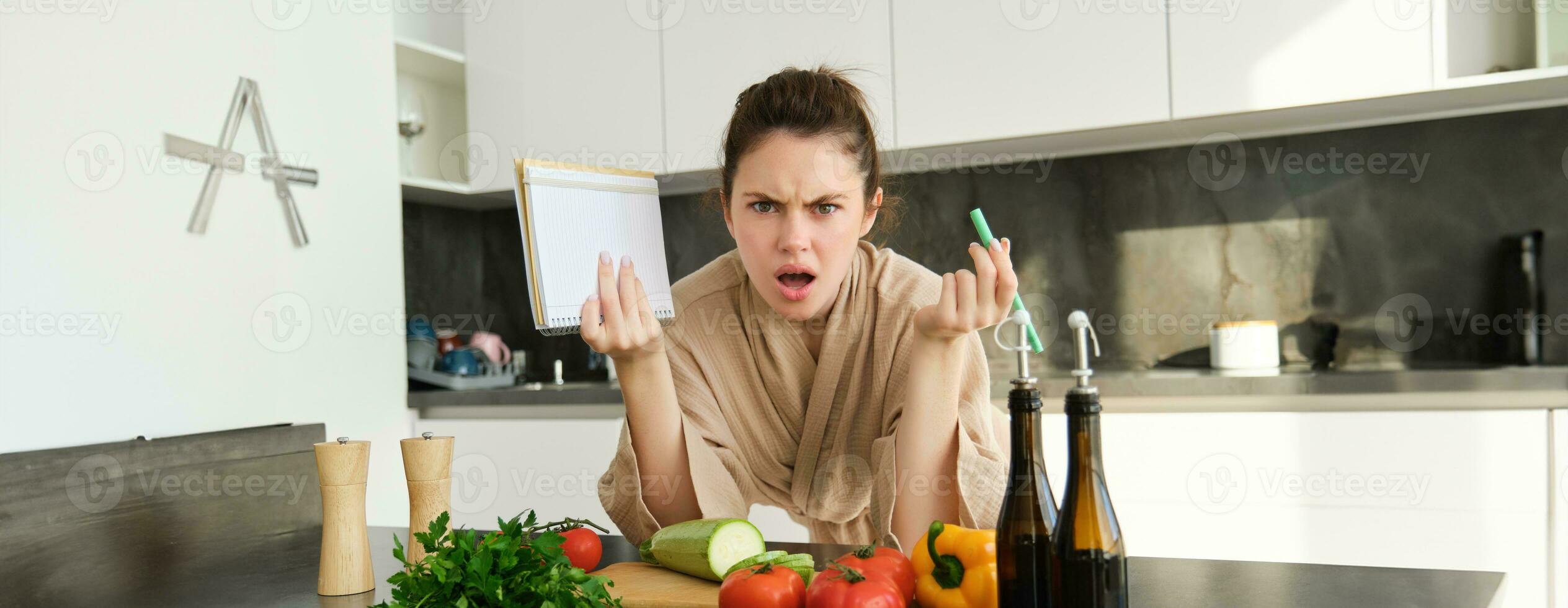 Portrait of woman with angry face, standing near vegetables and looking frustrated, holding notebook, annoyed while cooking meal photo