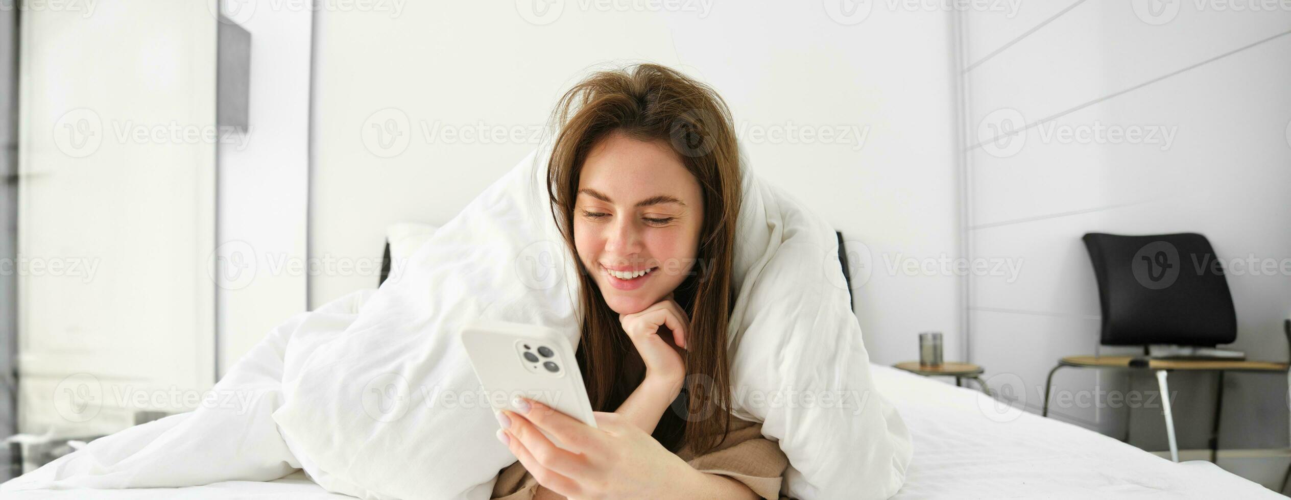Portrait of gorgeous girl wakes up in morning, lying in bed with smartphone, covered in blankets, reading social media feed on telephone, resting in bedroom photo