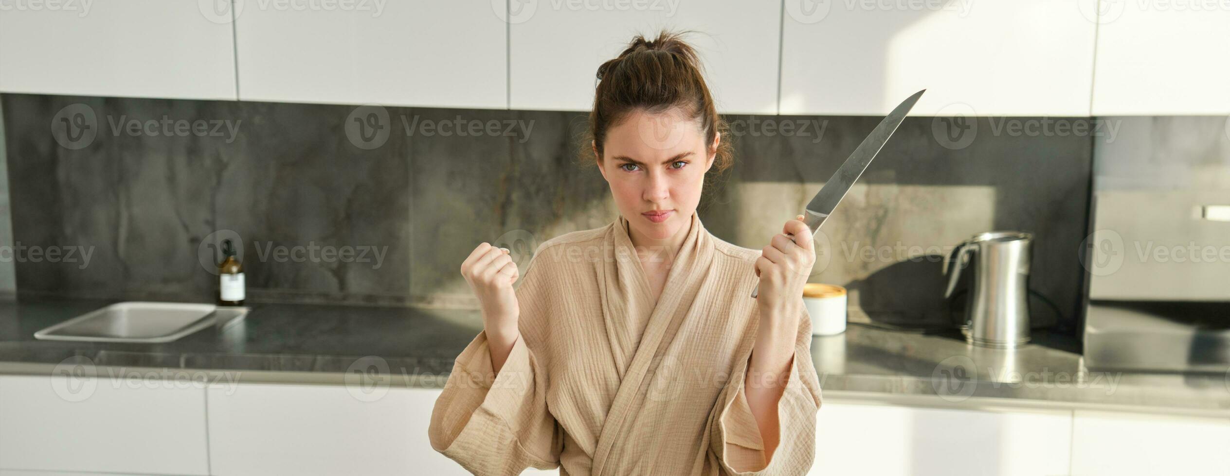 cerca arriba de grave y confidente morena mujer sostiene cuchillo y aprieta puño, en pie en el cocina, mirando seguro de sí mismo, posando en bata de baño foto