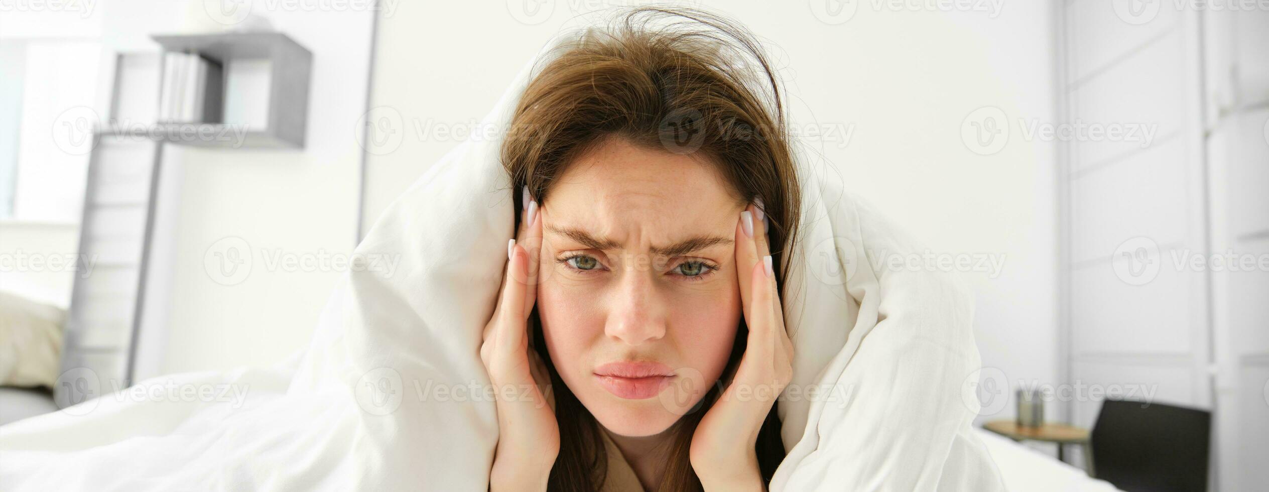 Woman feeling unwell in bed, lying in her bedroom under blanket and white sheets in morning, frowning and touching head, has headache or migraine photo