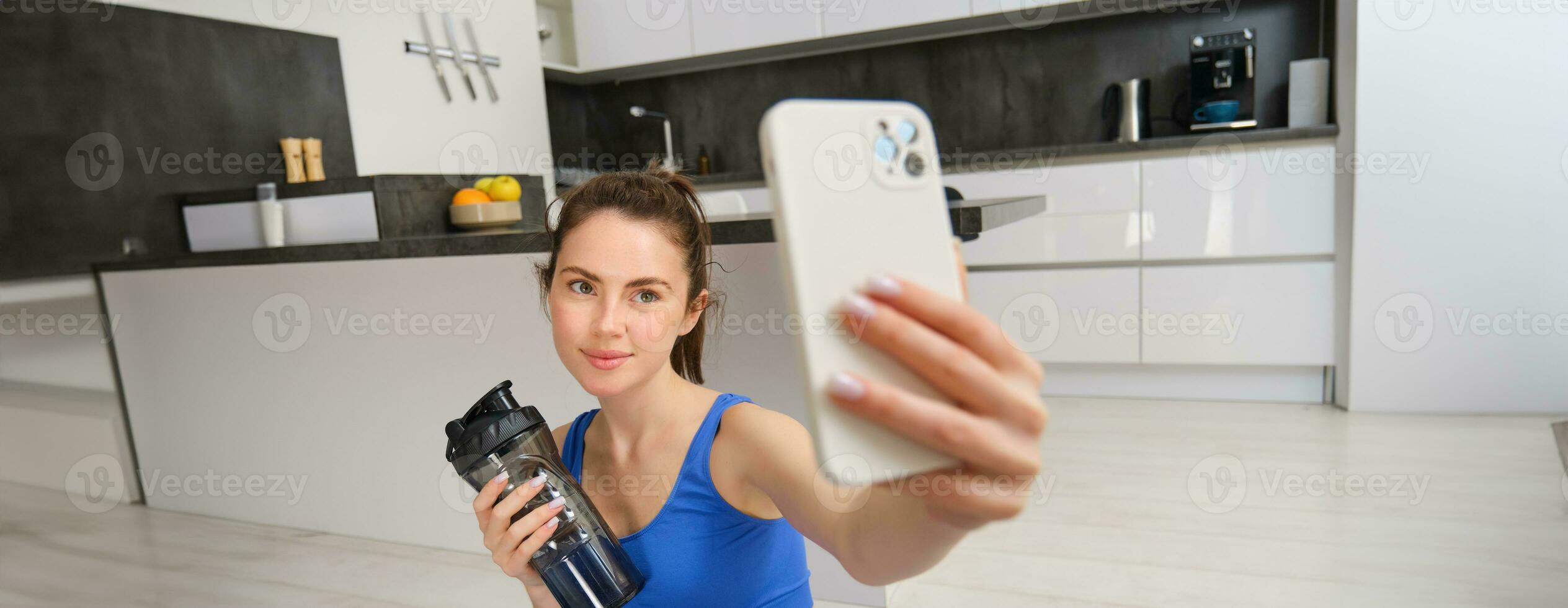 Portrait of young fitness girl, takes selfie on smartphone with water bottle, drinks water during workout, training at home, records sports vlog photo