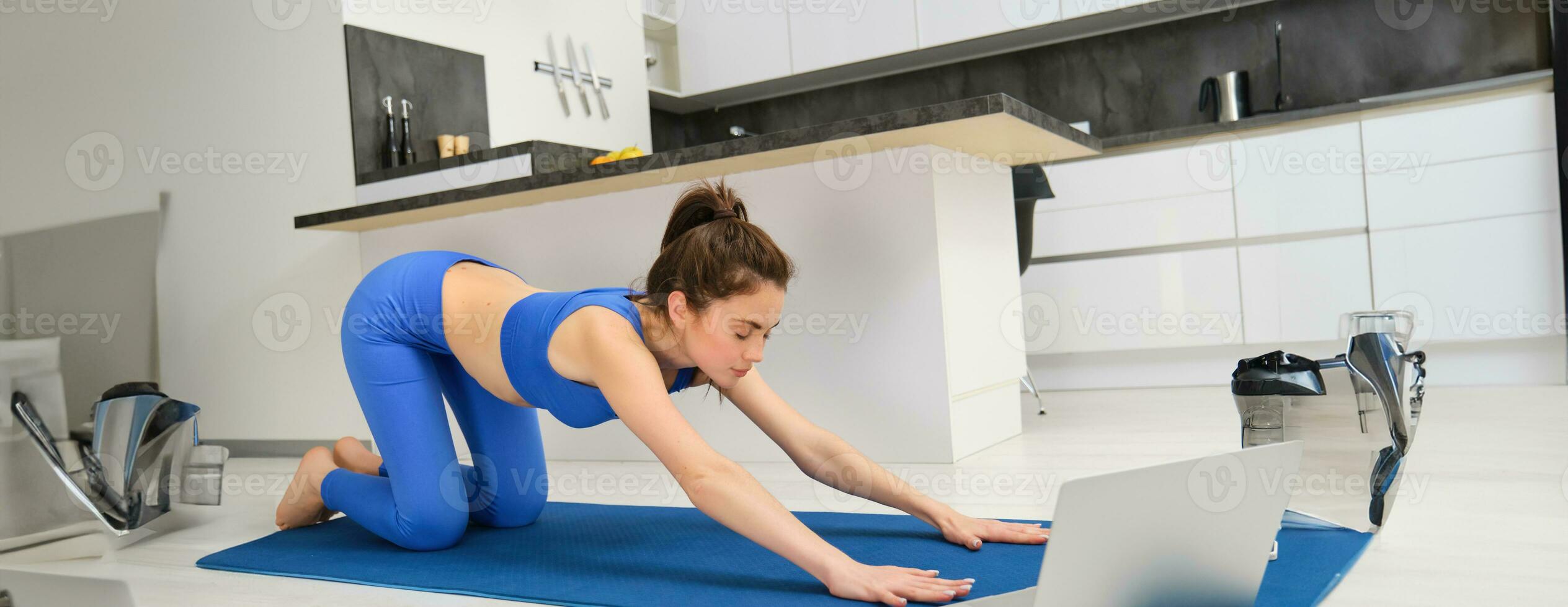 Portrait of woman doing stretching exercises, follows tutorial on laptop, attends online yoga class from home, workout on rubber mat photo