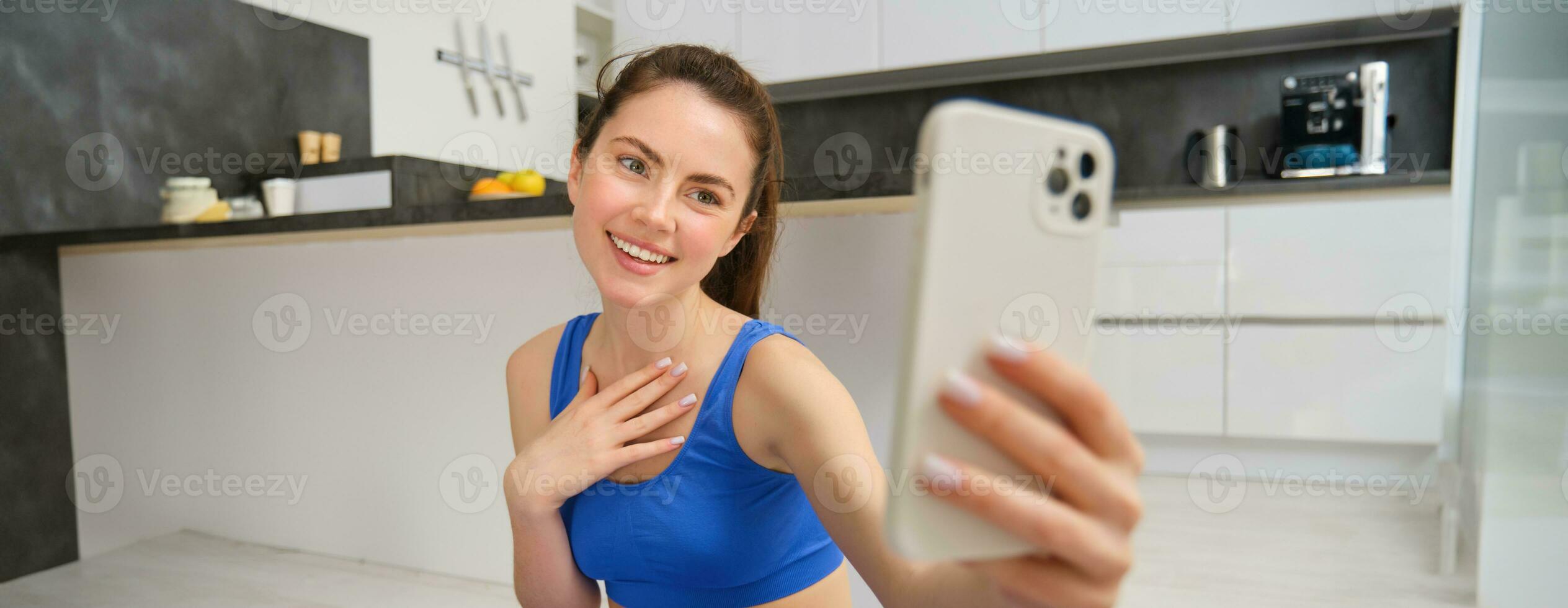 Smiling beautiful girl records video while doing sports at home, looks at smartphone, takes selfie on mobile phone, workout indoors in blue leggings and sportsbra photo