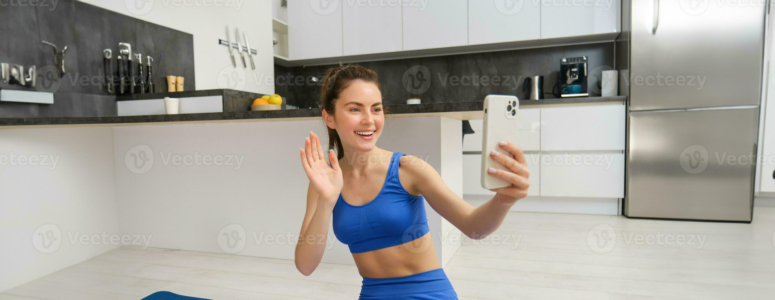 retrato de joven aptitud chica, blogger registros su rutina de ejercicio desde hogar, poses para selfie en teléfono inteligente, lo hace yoga en cámara, se sienta en azul sujetador deportivo y polainas foto