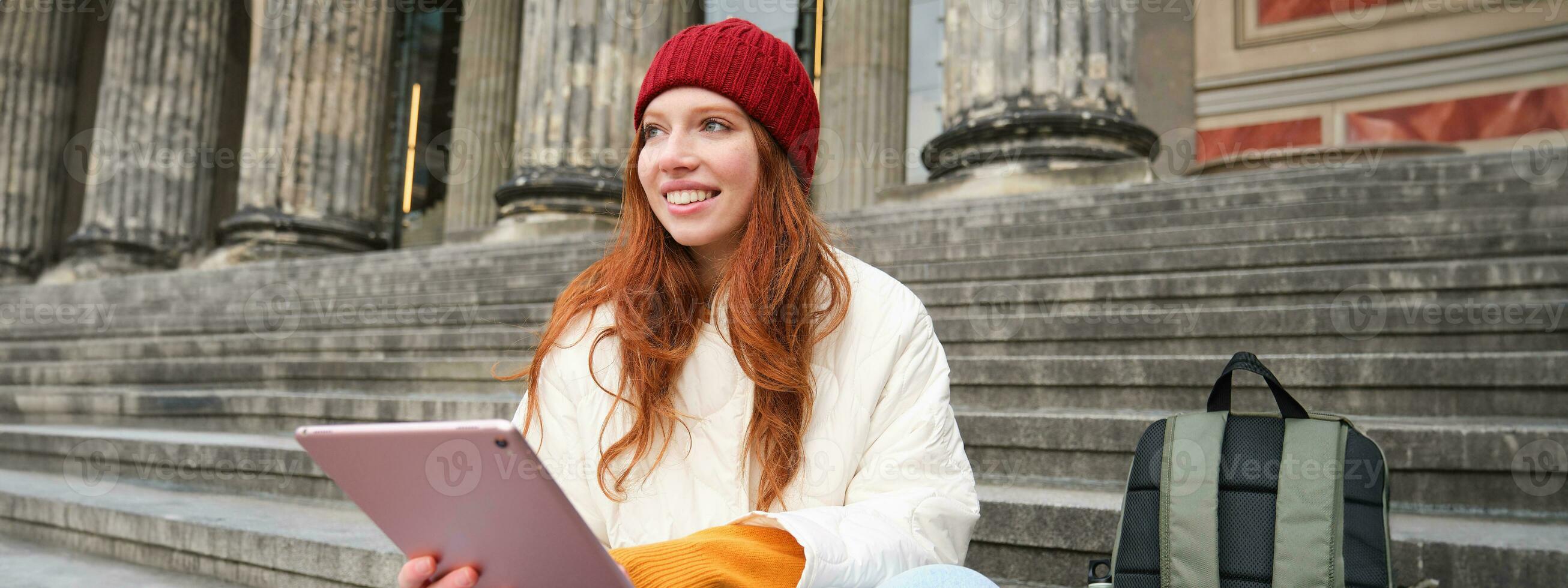 Beautiful young modern girl with red hair, holds digital tablet, sits on stairs near museum and connects public internet, sends message on gadget app photo