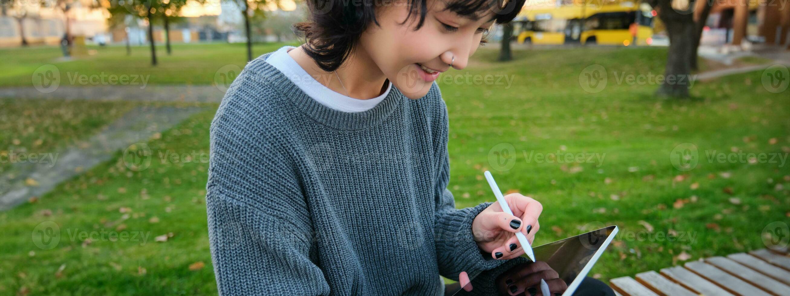 Portrait of young smiling korean girl, graphic designer, artist drawing on digital tablet with a pen tool, sitting in park on fresh air and scatching, taking notes photo