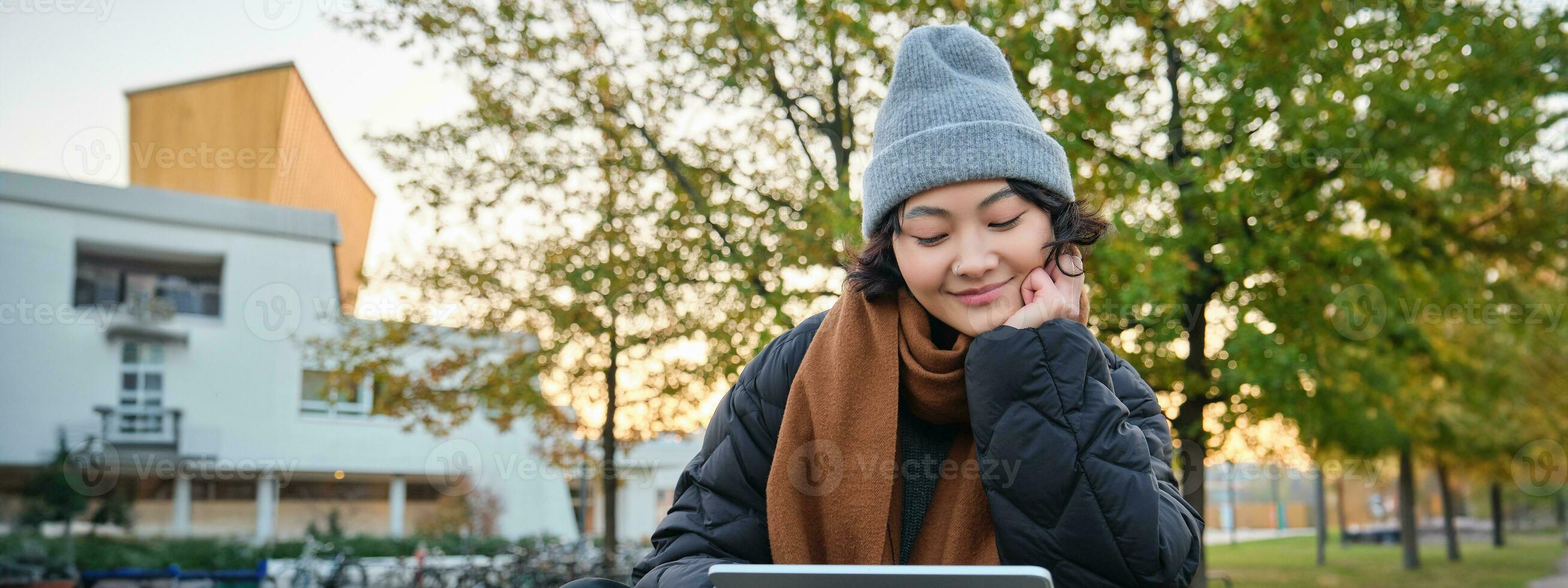Smiling asian girl, student sits on bench in park alone, reading, using social media app on digital tablet, watches videos outdoors, relaxes on fresh air photo