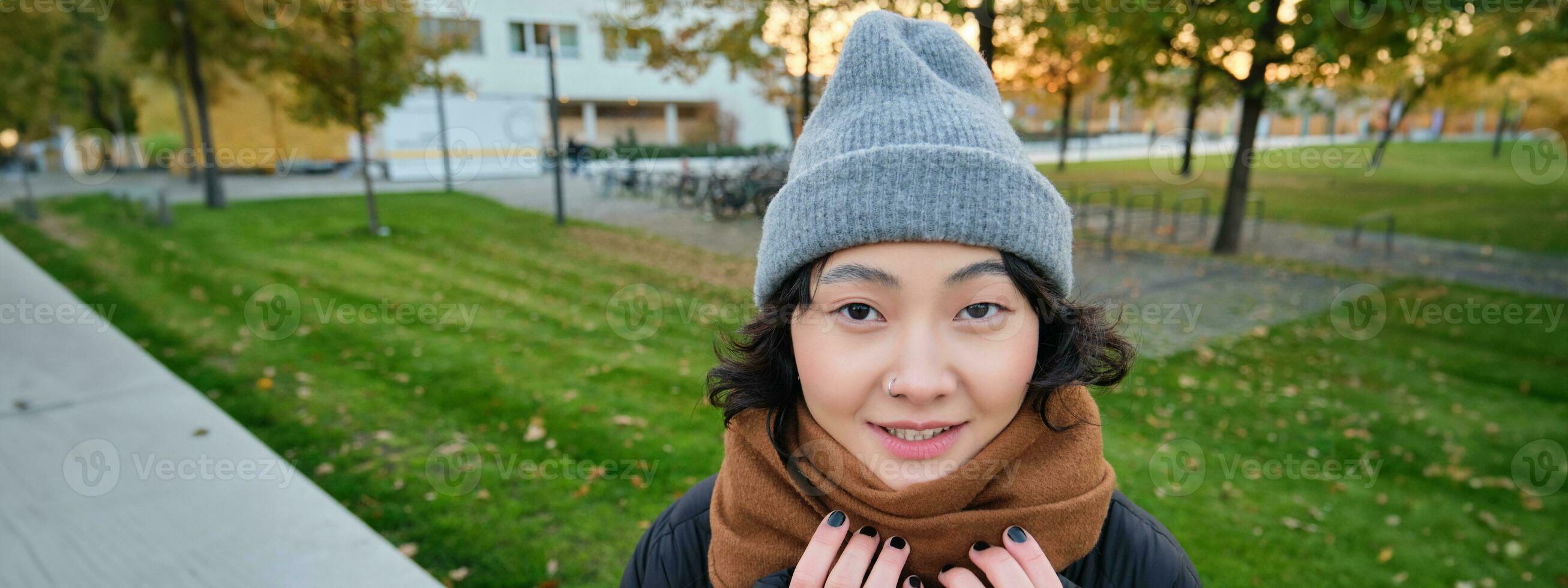 Portrait of cute asian girl in hat and scarf, walks around town in chilly spring weather, smiles and looks happy, sits in park near green grass photo